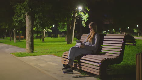 young woman sitting on park bench at night, deeply focused on her tablet under soft park lighting, surrounded by greenery and calm atmosphere, nighttime park setting