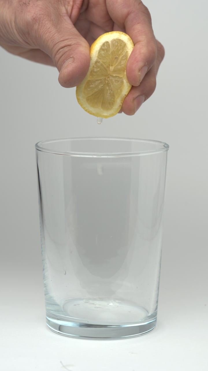 Vertical 9:16 slow motion video of a hand squeezing a fresh lemon as juice droplets fall into a transparent empty glass on a white background