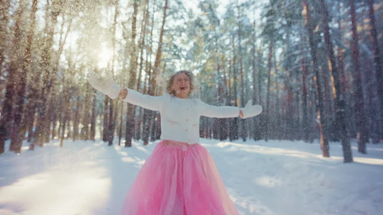 Ragazza gioiosa con un vestito rosa che gioca con la neve che cade in una foresta invernale.