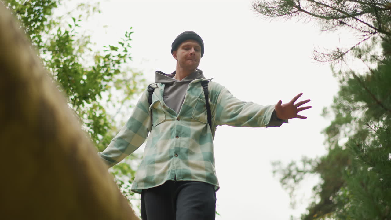 low angle forest white male hiker balancing on mossy log steadying arms with backpack, cautious expression, overcast sky, closeup perspective showing tension, balance challenge, travel grit