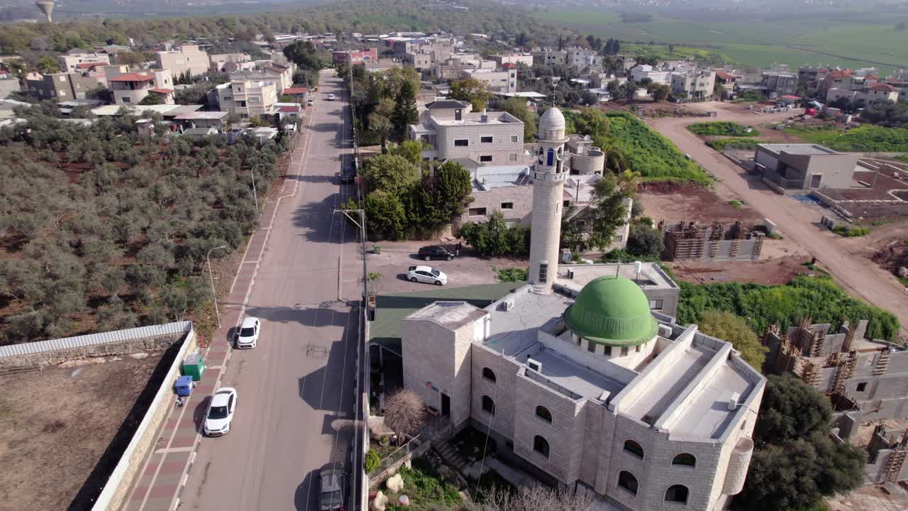 Aerial drone shot of a mosque in the Wadi Ara region of northern Israel, featuring a green dome and tall minaret as pigeons fly around the structure, with surrounding village buildings, roads, and agr