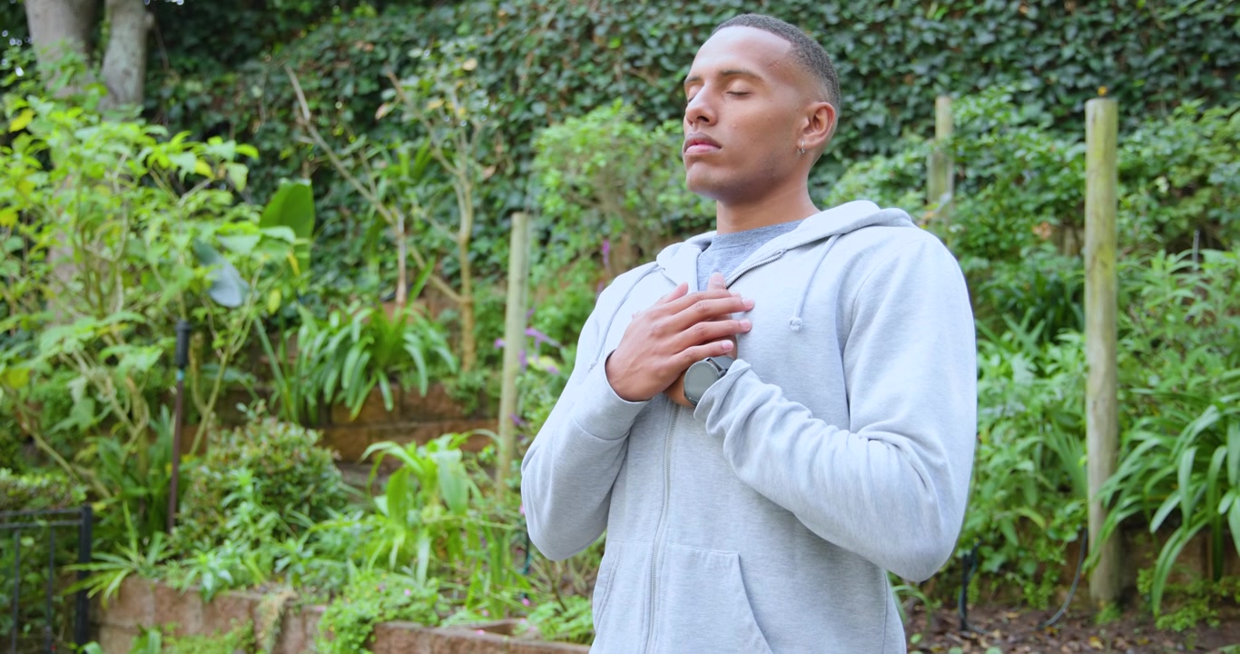 Un homme se préparant, les mains jointes et les yeux fermés, pratiquant une respiration apaisante dans un jardin, portant un sweat à capuche et une montre.