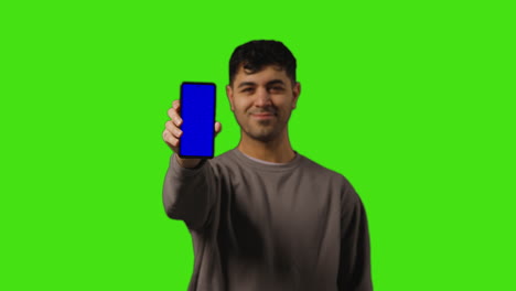 portrait of young man holding blue screen mobile phone to camera standing against green screen