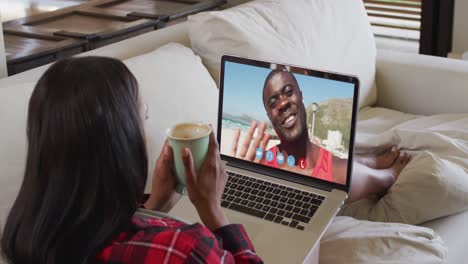 African-american-woman-holding-coffee-cup-having-video-call-on-laptop-while-sitting-on-couch-at-home