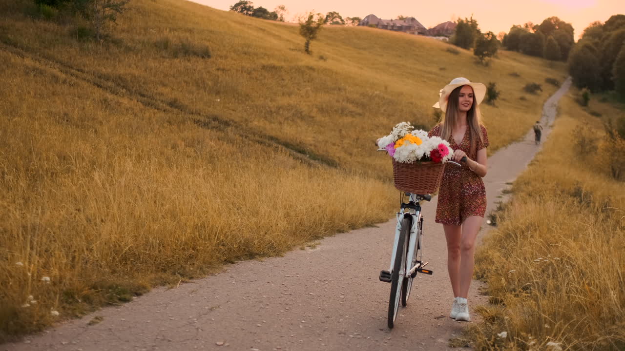 슬로우 모션 섹시한 아름다운 여성은 모자와 가운 여름 드레스를 입고 바구니에 꽃과 함께 와서 미소 짓고 있습니다.