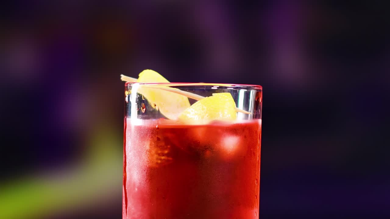 A vibrant red cocktail with an orange twist garnish against a blurred purple and green background.