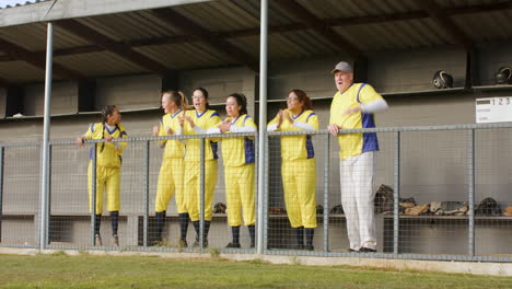 multiracial female baseball players and male coach watching the game and supporting on a pitch