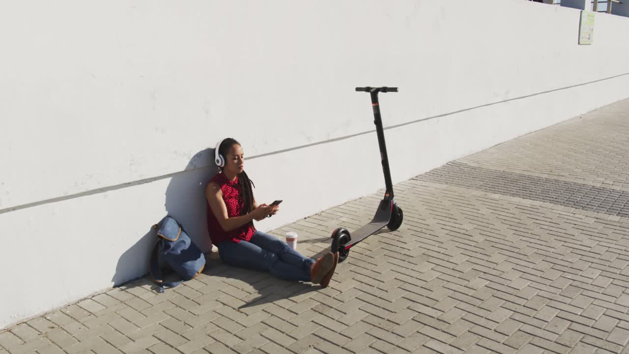 mujer afroamericana con auriculares usando un teléfono inteligente sentada en el pavimento con un scooter