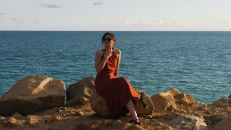 Woman-in-red-dress-sitting-on-rocky-coastline-at-Con-Dao-Island,-Vietnam-with-ocean-view