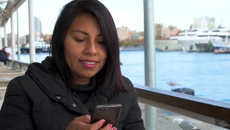 Latin-girl-sitting-on-a-terrace-by-the-sea-reads-messages-from-her-smartphone