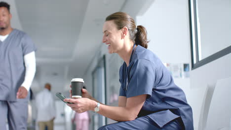 caucasian female doctor using smartphone and drinking coffee in corridor, copy space, slow motion