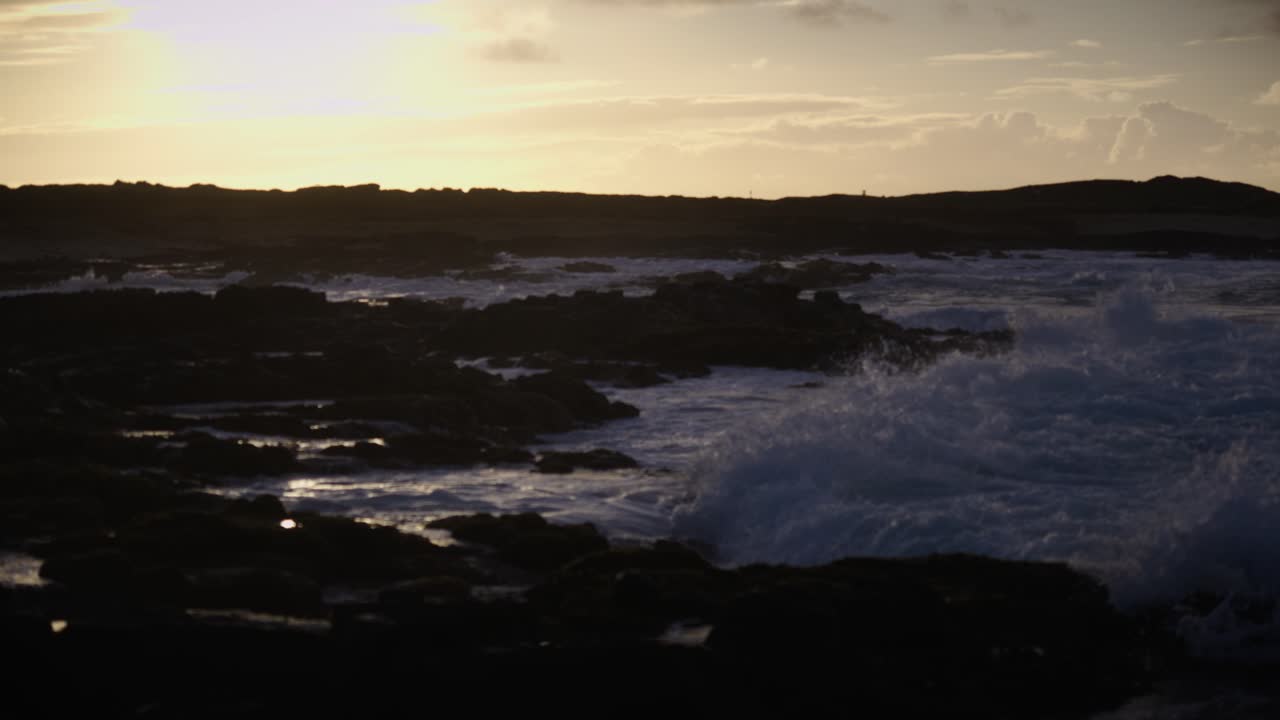 Powerful ocean waves crash into dark volcanic rocks as the setting sun bathes the coast in golden light, blending the raw force of nature with serene evening calm