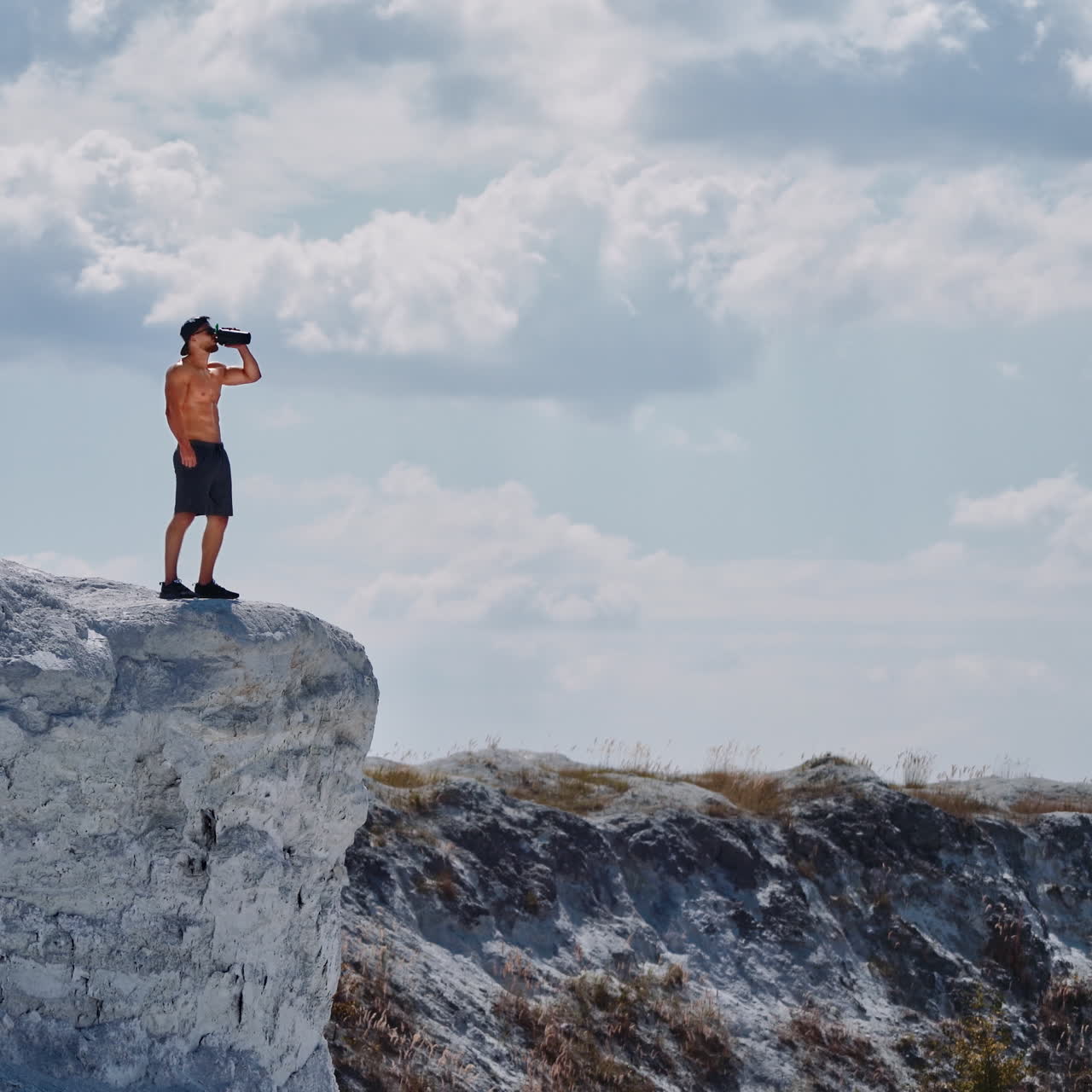 Athlete drinking water on a white rock in summer. Shirtless man with flask in white canyon in a sunny day. Healthy sportsman after training outdoors.