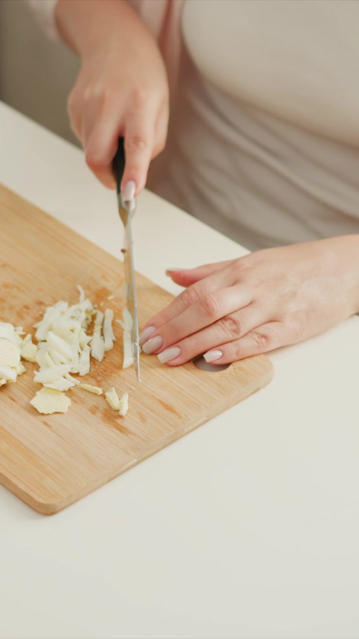 Una donna affetta con cura un uovo sodo su un tagliere di legno, dimostrando precise abilità con il coltello e nella preparazione del cibo. Le sue mani sono ferme mentre taglia la parte bianca a piccoli pezzi.