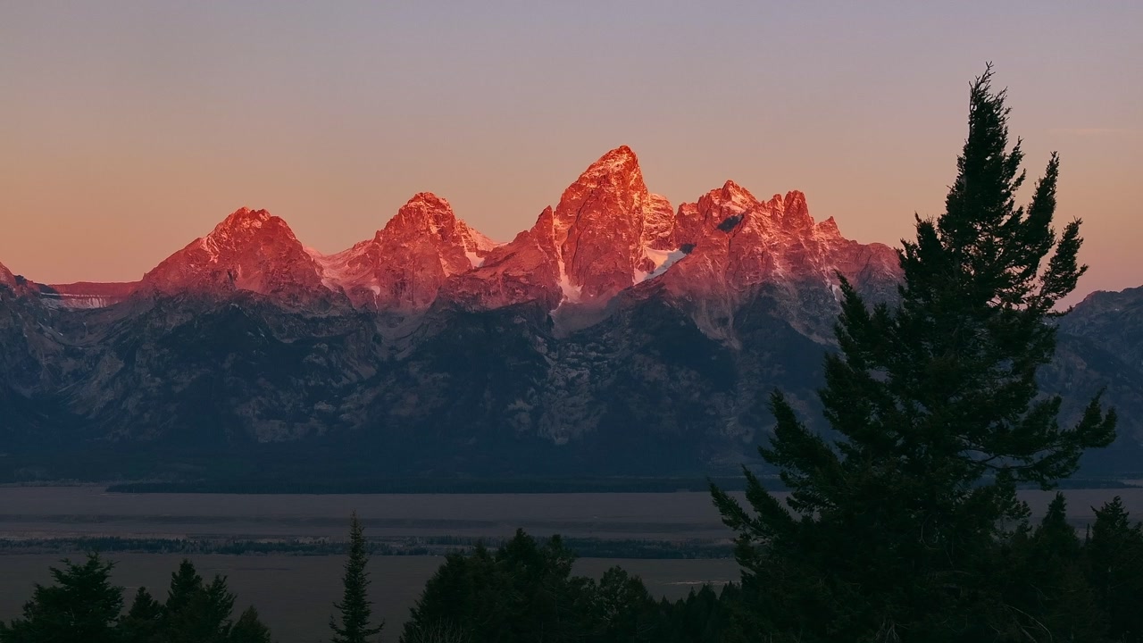 Snow capped Grand Teton peaks glowing in sunrise above forest silhouette, golden hour stunning backdrop for adventure