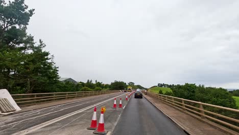 a vehicle travels along a rural english road undergoing maintenance, passing traffic cones, signage, construction equipment, and temporary barriers under overcast daylight