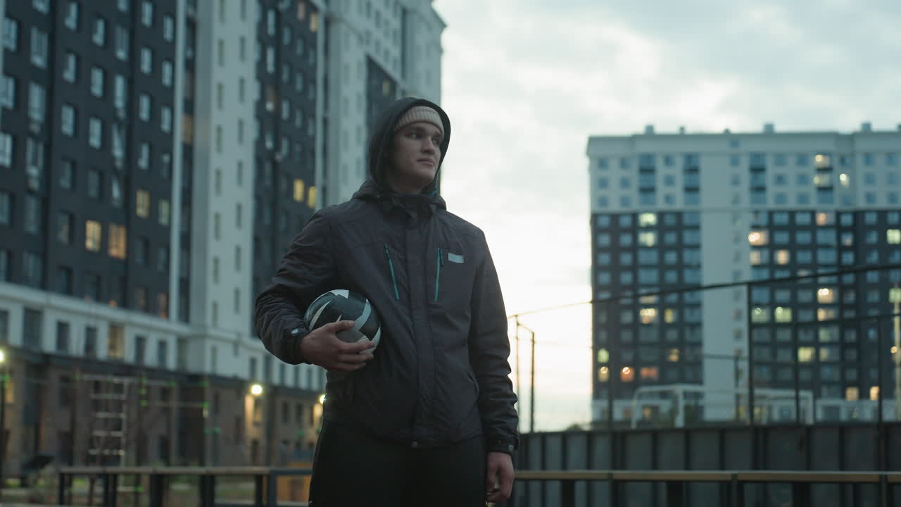 Athlete holding soccer ball looking tired during urban training session, standing confidently in modern sport arena surrounded by tall residential buildings