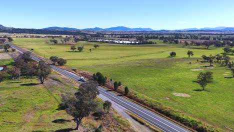 drone footage glides above a rural road winding through green fields, scattered trees, and distant hills under bright midday sunlight in new south wales