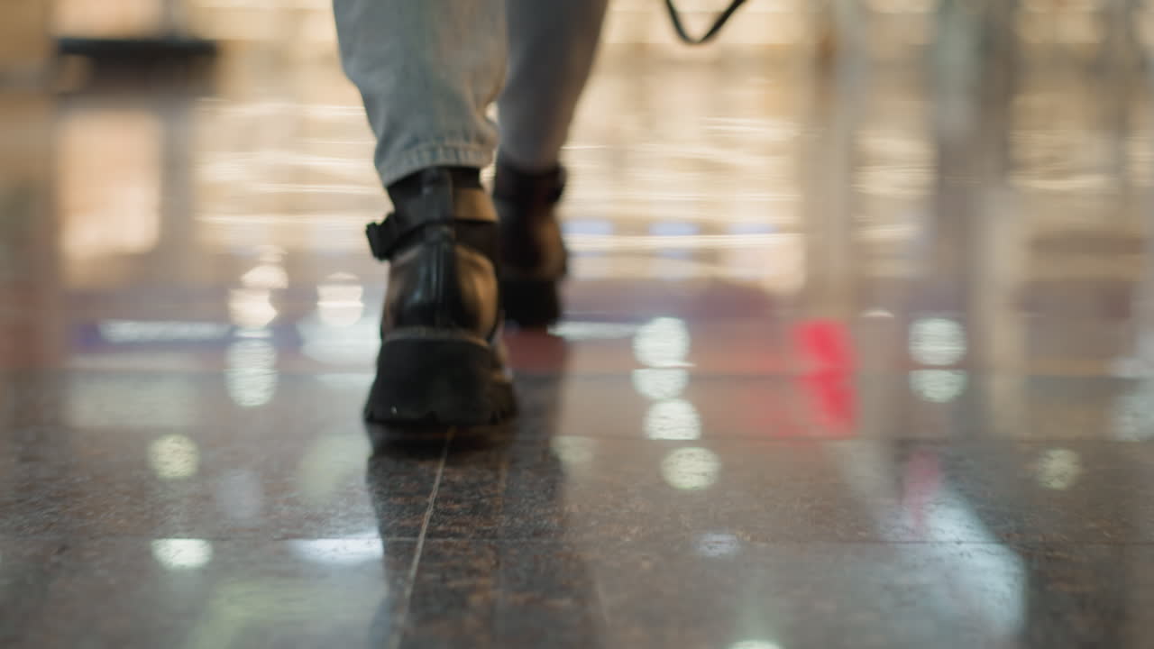 close up leg view of woman wearing chunky black boots walking on glossy tiled floor under bright overhead lights with reflection capturing motion and style in modern mall corridor retail interior