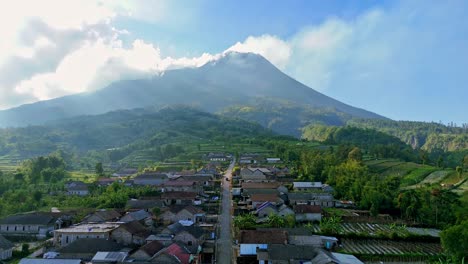 drone fly over village house located on mountain slope with scenery of active merapi volcano emitting smoke from the peak and cloudy blue sky