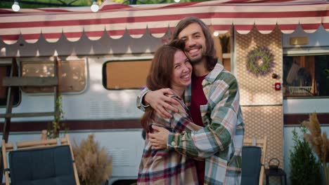 Portrait-of-a-happy-brunette-girl-in-a-pink-checkered-shirt-who-runs-to-her-brunette-boyfriend-in-a-green-checkered-shirt-and-hugs-him-in-their-trailer-while-relaxing-in-a-camp-during-a-picnic-in-the-summer