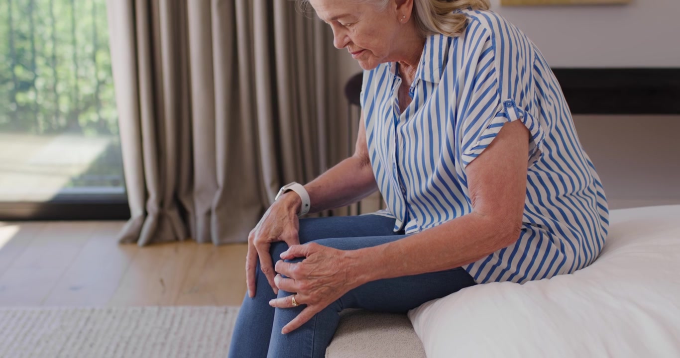 Donna anziana seduta in camera da letto, con una blusa a righe e jeans, che si strofina le ginocchia preparandosi ad alzarsi.