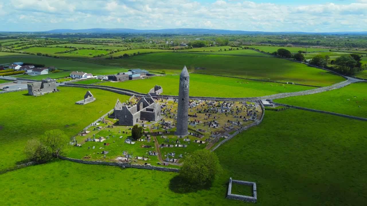 Aerial spin shot of Kilmacduagh abbey ruins with patches of sun on surrounding fields and grasslands.