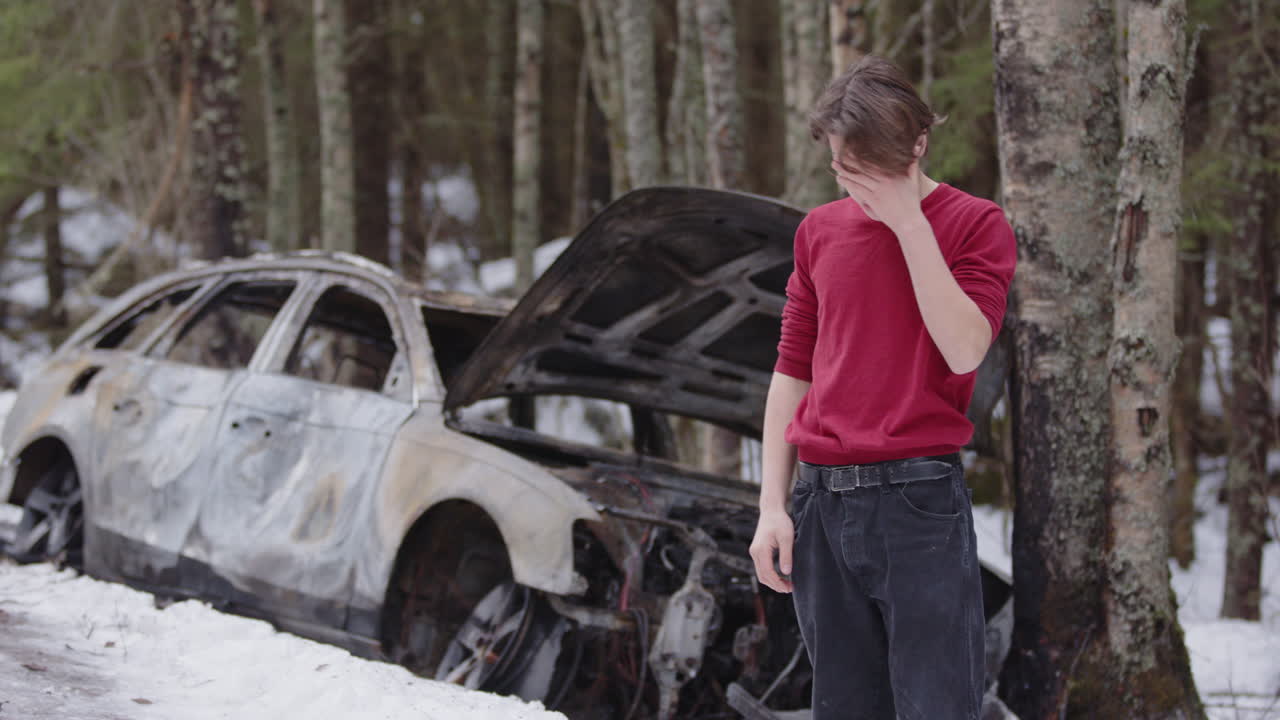 Very emotional young man stands near wrecked and burnt out car on snowy roadside