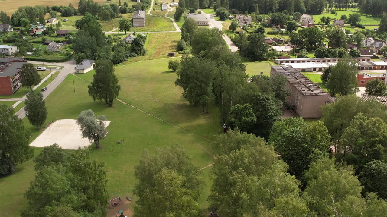 Aerial view of a small town hosting a lively outdoor fair with market stalls, parked cars, houses, and surrounding green fields under a sunny sky. Jaunpiebalga