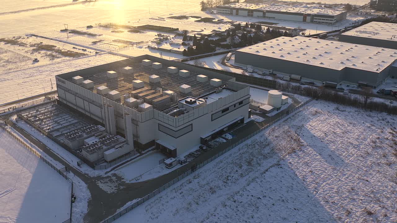 Establishing shot of a large, modern data center facility during a cold winter sunrise. The surrounding landscape is covered in snow