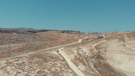 Drone-shot-in-the-Judean-desert,-brown-hills,-sandy-path,-cars-driving-on-the-road,-electrical-poles-and-small-towns-far-away,-Israel