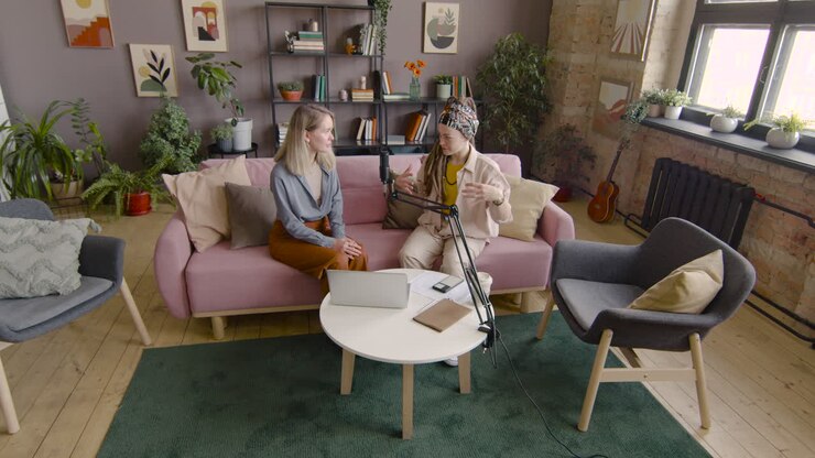 Zoom In And Top View Of Two Women Recording A Podcast Talking Into A Microphone While Sitting On Sofa In Front Of Table With Laptop And Documents