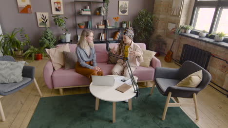 Zoom-In-And-Top-View-Of-Two-Women-Recording-A-Podcast-Talking-Into-A-Microphone-While-Sitting-On-Sofa-In-Front-Of-Table-With-Laptop-And-Documents