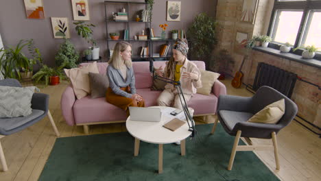 zoom in and top view of two women recording a podcast talking into a microphone while sitting on sofa in front of table with laptop and documents