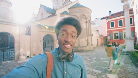 hombre afroamericano sosteniendo un teléfono inteligente y hablando con la cámara mientras señala a sus amigos detrás
