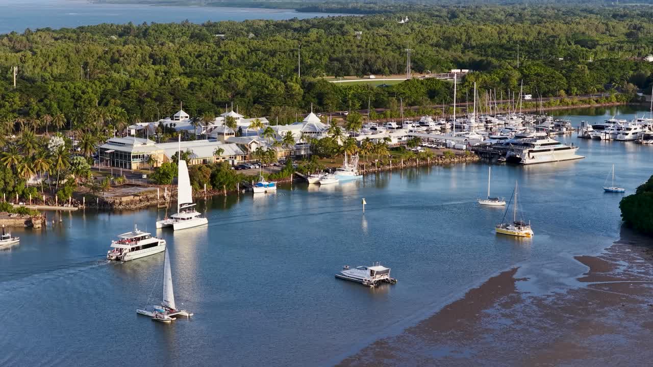 Drone footage captures boats navigating a tropical marina in Port Douglas, Australia, under clear skies with lush greenery surrounding