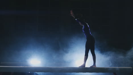 la niña realiza un truco en un tronco con luz de fondo y cámara lenta en ropa de gimnasia deportiva. humo y azul. saltar y girar en la viga de equilibrio