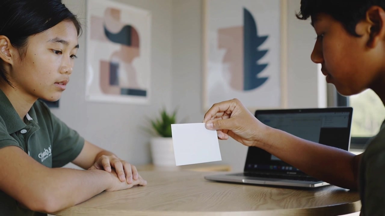 Bringing right teen showing white card to left teen at home with laptop, wearing green shirts