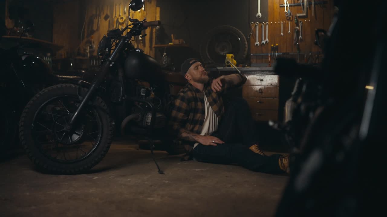 은 셔츠를 입은 피곤한 남자 메카닉이 이마를 고 오토바이에 기대어 그의 워크 스튜디오에서 휴식을 취합니다.
