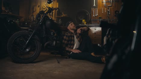 vista lateral de un mecánico cansado en una camisa a cuadros se limpia la frente y se inclina sobre una motocicleta para descansar en su estudio de taller