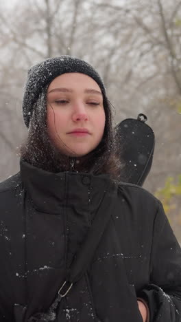 white chic in winter jacket adjusts her guitar in its pack, gazing thoughtfully into the distance, with a snow-covered bench in the background amidst falling snow