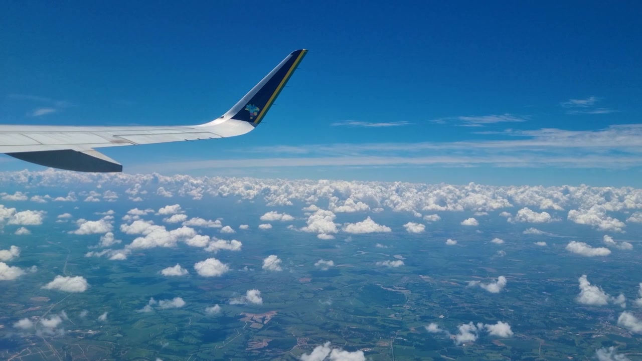 Flyfoto fra et flyvindu over Paraná, Brasil, med spredte skyer som driver over enorme grønne jordbruksområder og landlige landskap under en lys blå himmel, som fanger reisens skjønnhet