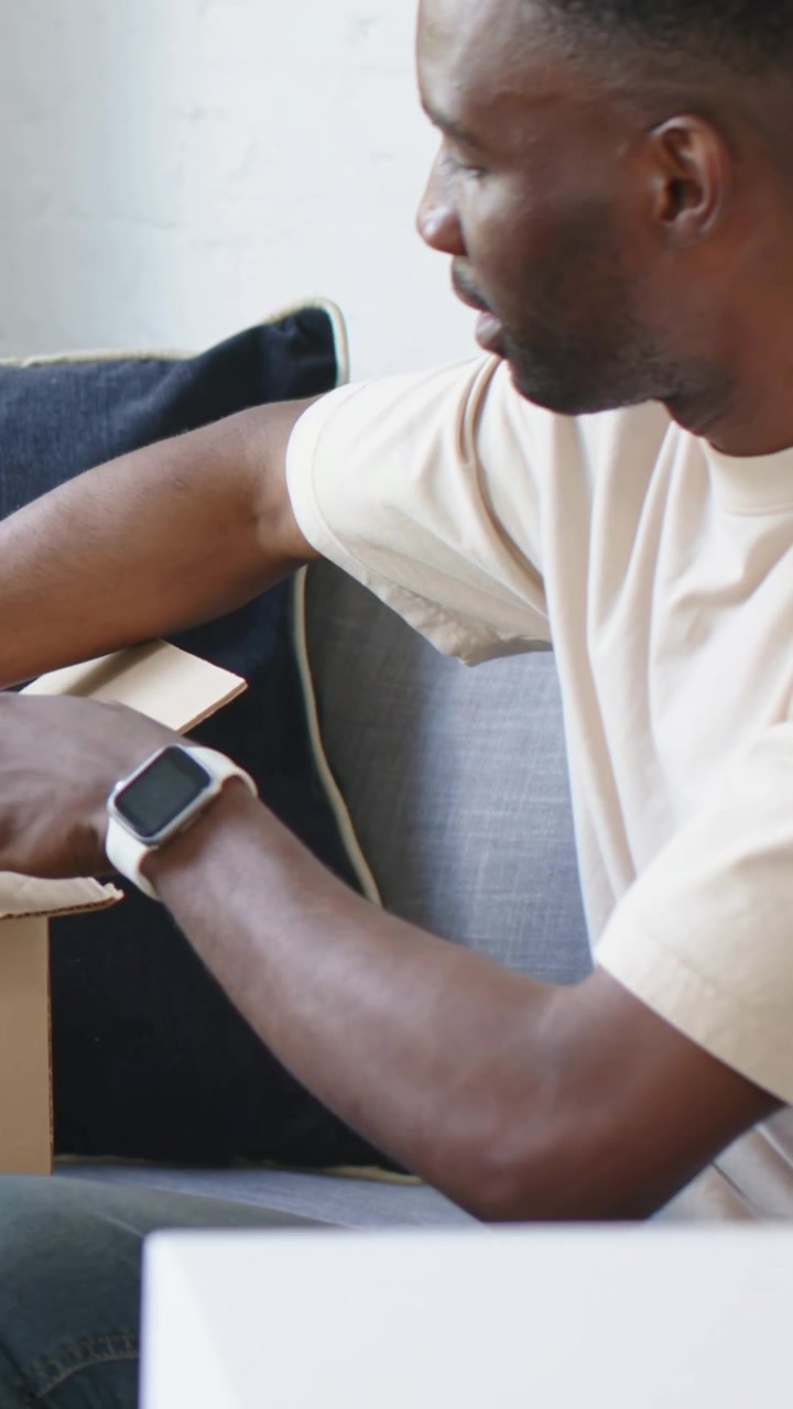 Vertical video: Leaning man in tee, jeans opening, checking box on couch at home watch, copy space