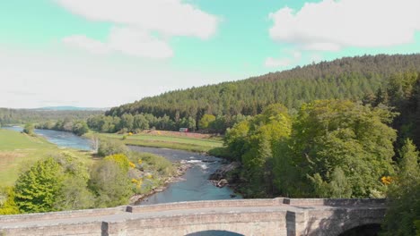 Busfahrt-Entlang-Des-Flusses-Mit-Brücke-Im-Sonnigen-Schottischen-Hochland,-Drohne