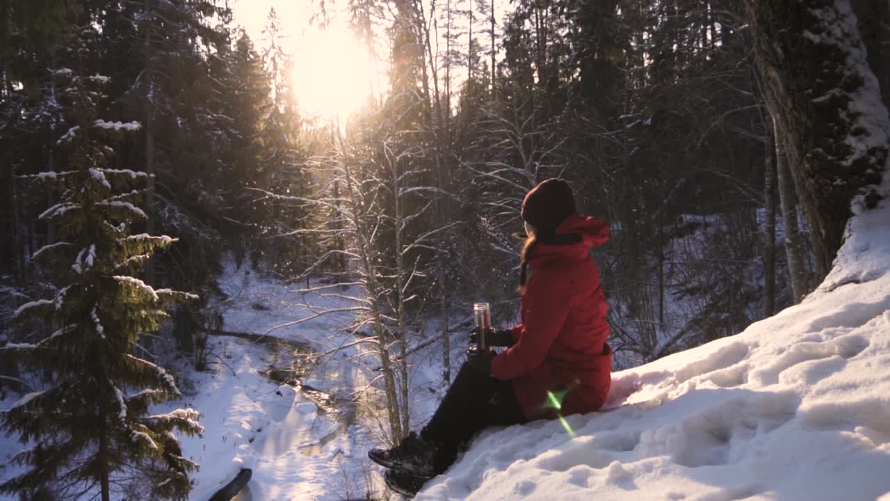 chica con abrigo de invierno rojo sentada en una colina nevada sosteniendo un vaso de té y admirando la brillante luz del sol