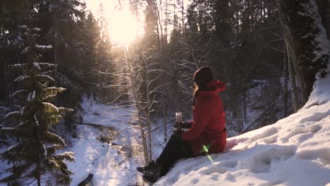 girl in red winter coat sitting on snowy hill holding a glass of tea bottle and admire the bright sunlight