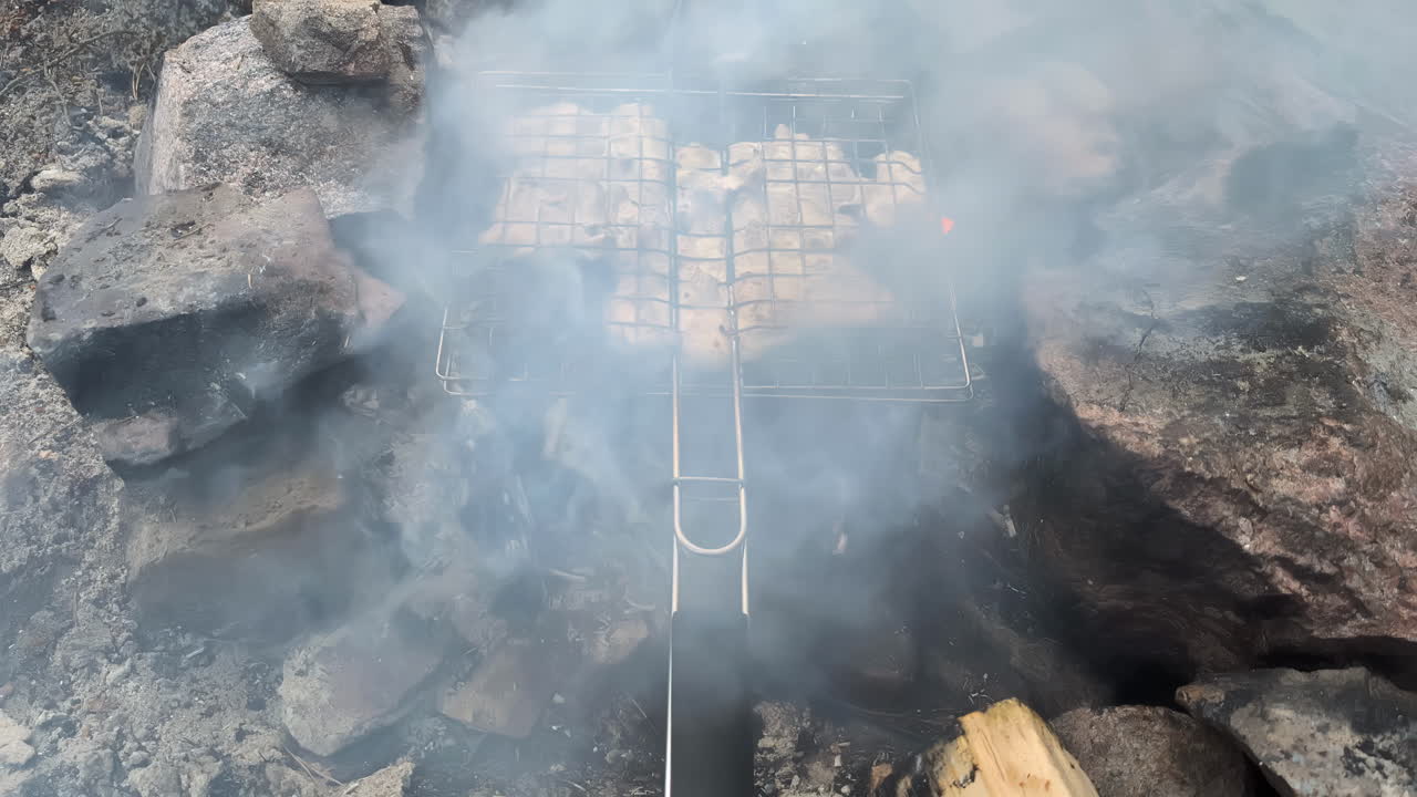Close-up of meat chicken food slow cooking on outdoor fire kitchen utensil grill surrounded by rocks with heat warmth and smoke burning embers charcoal campfire