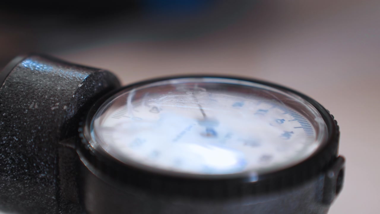 Manometro della pressione sanguigna in primo piano su bracciale nero, paramedico a domicilio valuta paziente anziano durante chiamata urgente, messa a fuoco superficiale, luce ambientale soffusa, quadrante usurato, sfocatura di movimento, atmosfera tesa, attrezzatura portatile.