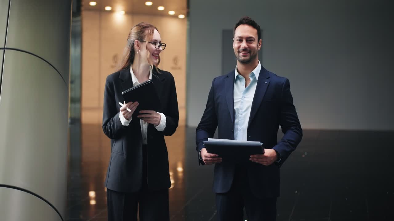Two business professionals meeting in dark hallway with documents and portfolio in hand, laughing as they prepare for meeting