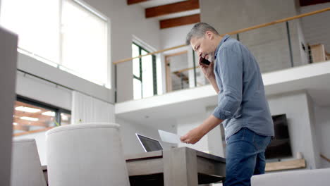 senior biracial man on phone, standing looking at paperwork, home working, copy space, slow motion